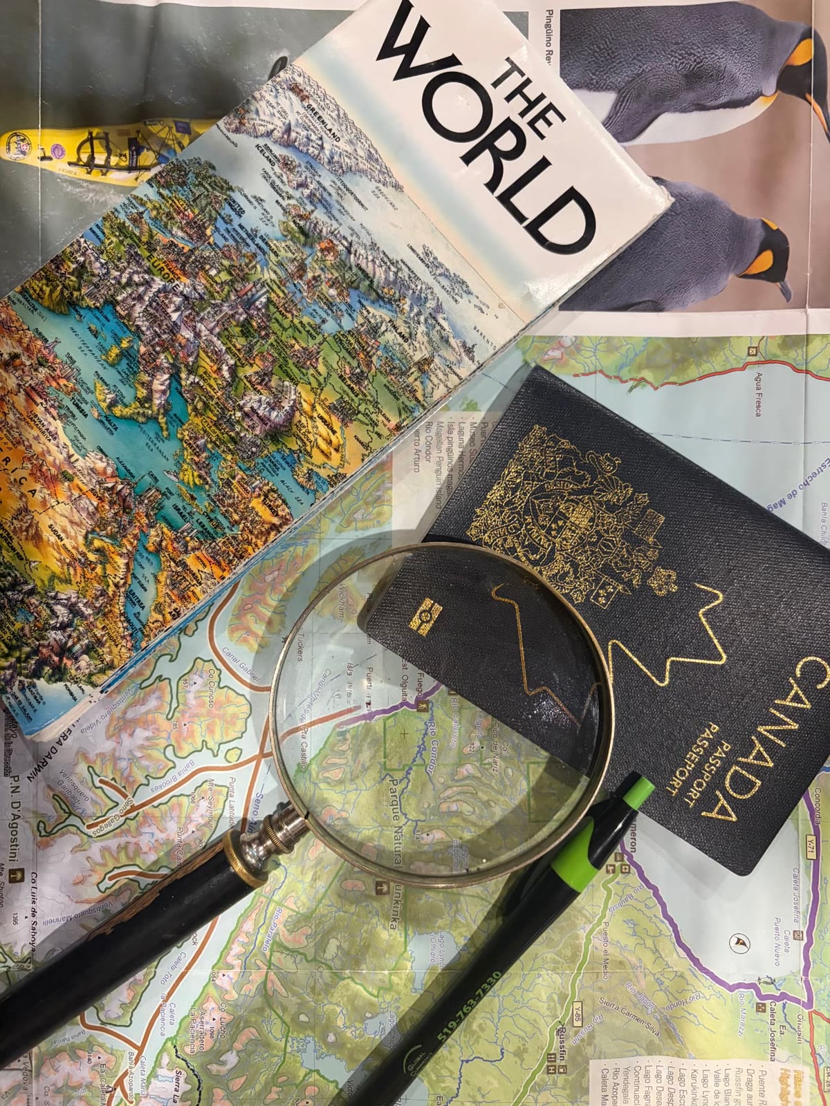 A flat lay of travel planning essentials: world maps, a Canadian passport, a magnifying glass and a pen